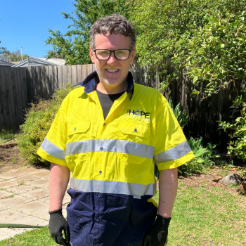 A smiling man wearing glasses and a Hope Industries yellow high-visibility shirt standing in a sunny backyard garden.